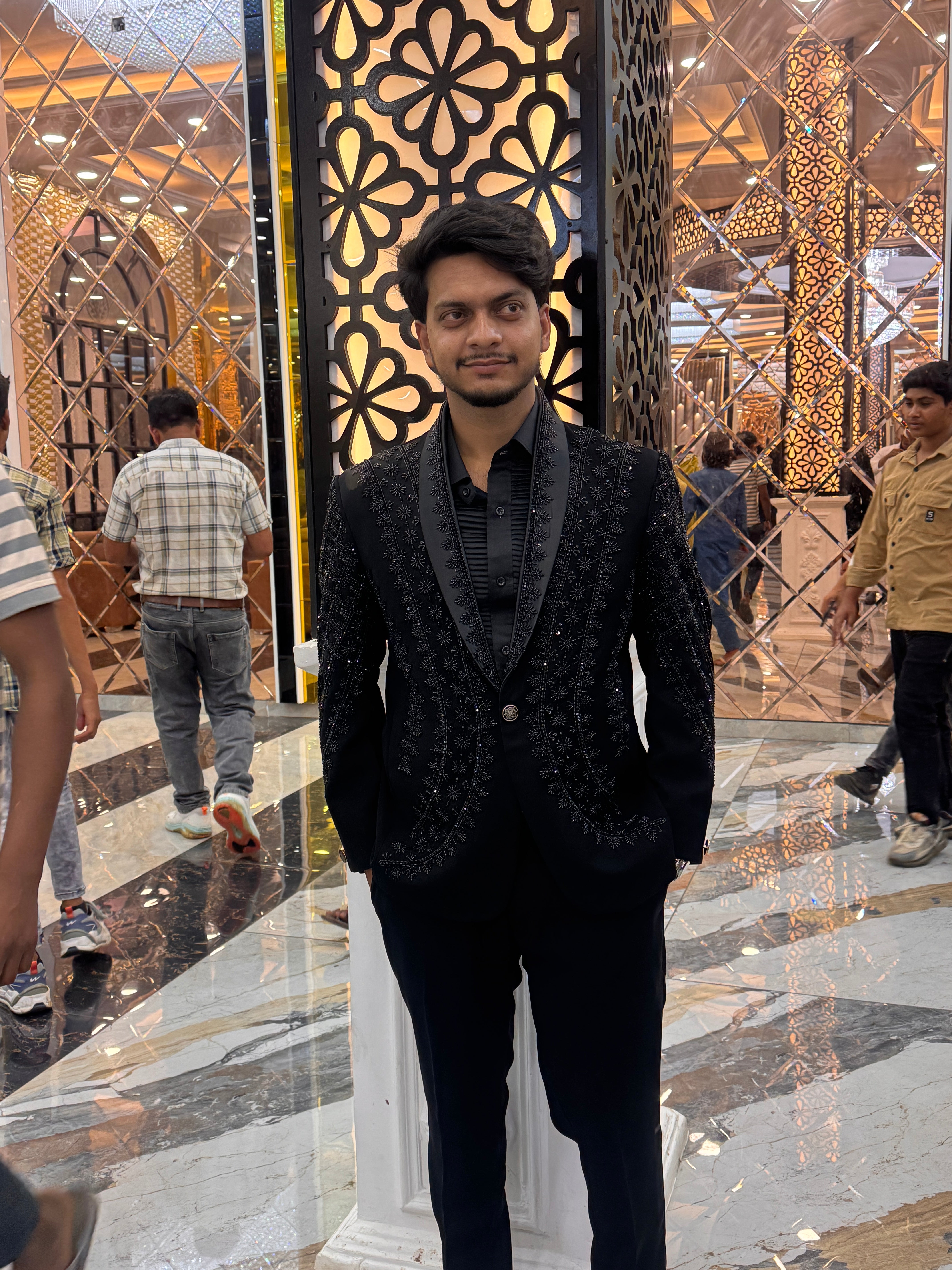 A man in a white, hand-painted tuxedo suit with colorful floral and cactus-like designs. He is sitting on a chair with one arm resting on the side, looking at the camera.

